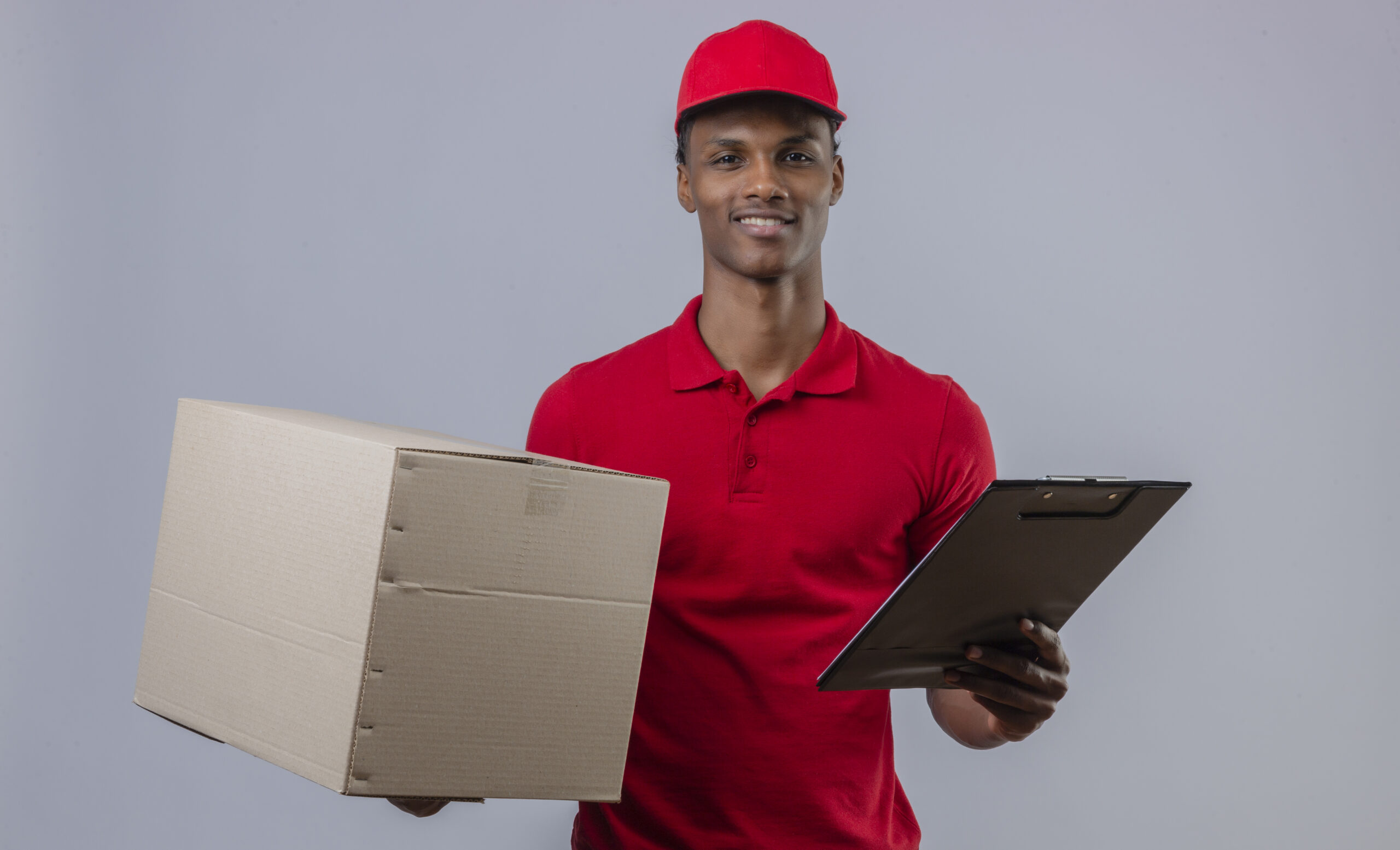 young african american delivery man wearing red polo shirt and cap holding cardboard box and clipboard with smile on face standing over isolated white background